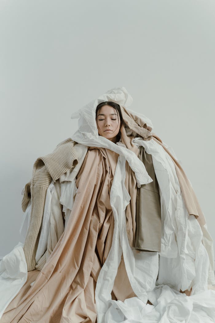 A woman surrounded by a pile of assorted neutral colored clothes, showcasing consumerism excess.